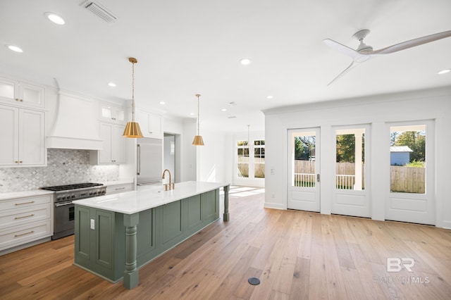 kitchen featuring a kitchen island with sink, premium range hood, stainless steel stove, decorative light fixtures, and white cabinetry