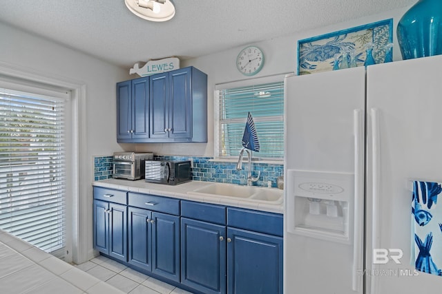 kitchen with tile countertops, blue cabinets, sink, white fridge with ice dispenser, and a textured ceiling