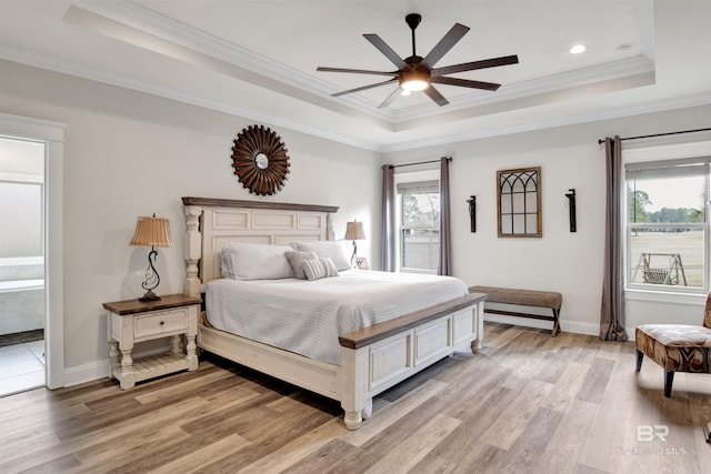 bedroom featuring connected bathroom, light wood-type flooring, a tray ceiling, ceiling fan, and multiple windows