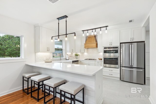 kitchen featuring white cabinets, kitchen peninsula, hanging light fixtures, custom range hood, and stainless steel appliances