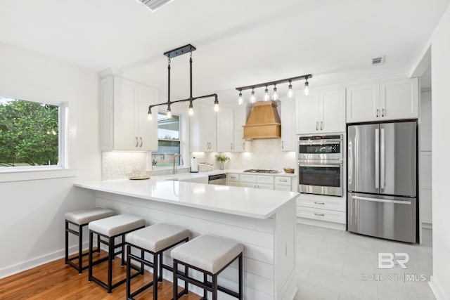 kitchen with light countertops, custom exhaust hood, a peninsula, appliances with stainless steel finishes, and backsplash