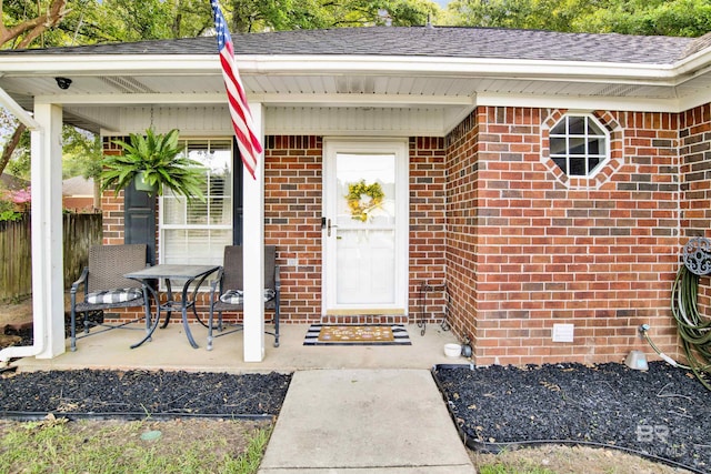 property entrance featuring brick siding, a porch, a shingled roof, and fence