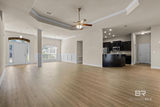 unfurnished living room with decorative columns, a tray ceiling, ornamental molding, light hardwood / wood-style floors, and ceiling fan with notable chandelier