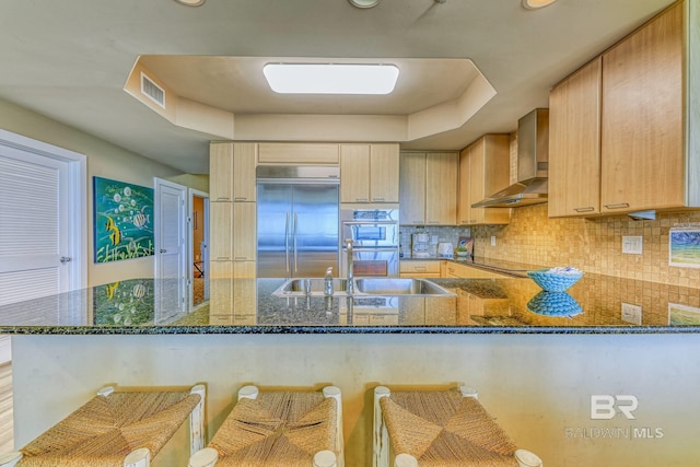 kitchen with a breakfast bar area, appliances with stainless steel finishes, wall chimney range hood, and dark stone countertops