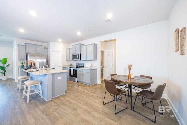 kitchen with an island with sink, stainless steel appliances, a breakfast bar area, light stone countertops, and light hardwood / wood-style floors