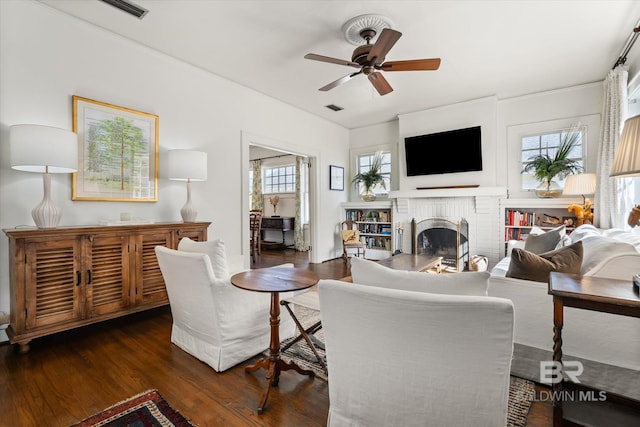 living room with a brick fireplace, wood finished floors, visible vents, and ceiling fan