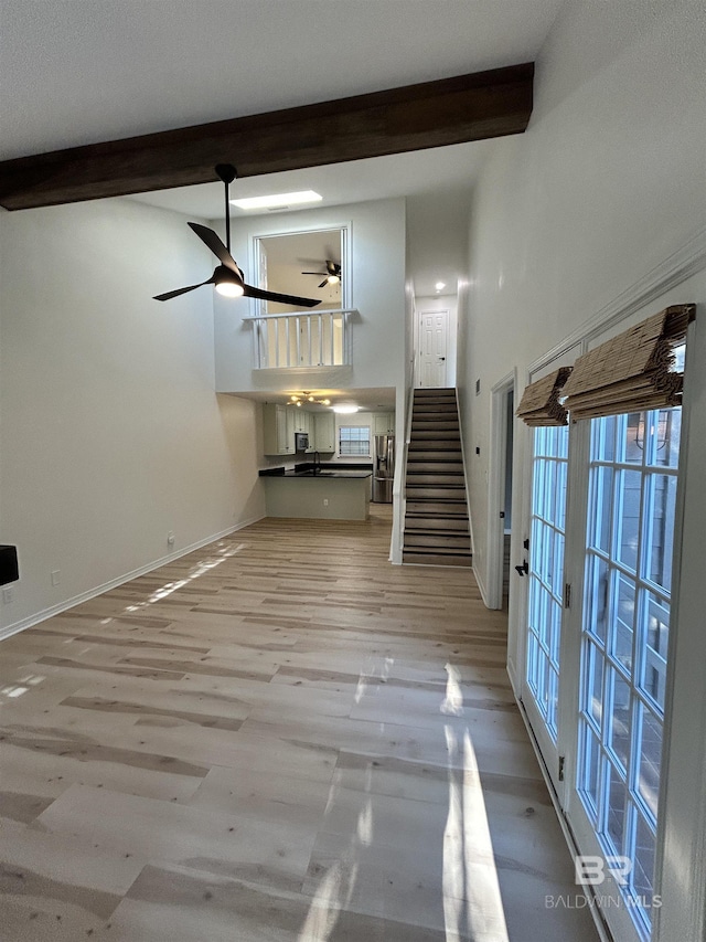 unfurnished living room featuring stairway, a ceiling fan, light wood-style floors, beamed ceiling, and baseboards