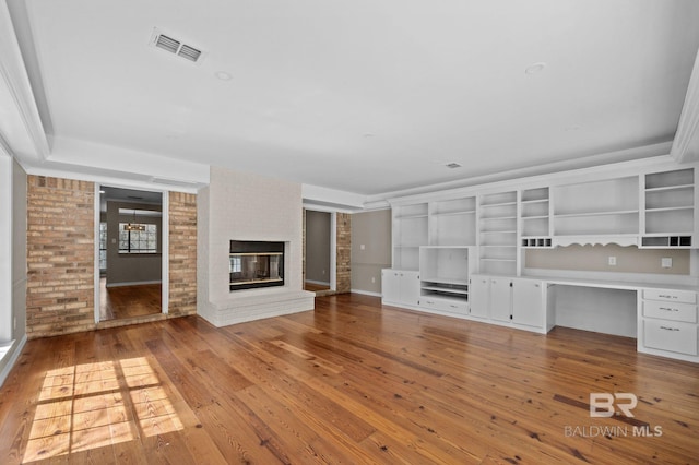 unfurnished living room with a brick fireplace, built in study area, visible vents, and wood-type flooring