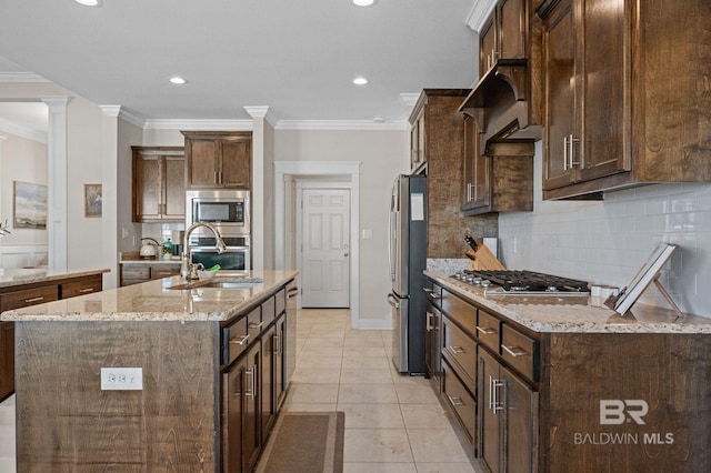 kitchen with sink, stainless steel appliances, light stone counters, decorative columns, and an island with sink