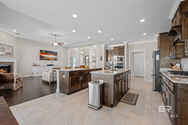 kitchen featuring a kitchen island with sink, ornamental molding, stainless steel appliances, and a brick fireplace