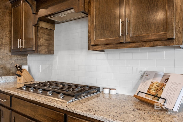 kitchen featuring light stone countertops, tasteful backsplash, custom range hood, dark brown cabinets, and stainless steel gas cooktop