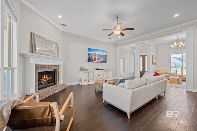 living room featuring dark wood-type flooring, decorative columns, a fireplace, ceiling fan with notable chandelier, and ornamental molding