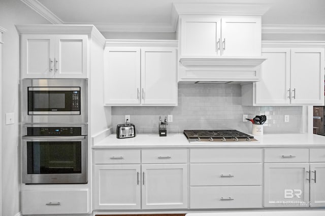 kitchen with white cabinets, appliances with stainless steel finishes, ornamental molding, tasteful backsplash, and custom range hood