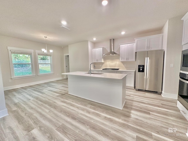 kitchen with sink, wall chimney range hood, stainless steel appliances, white cabinets, and a kitchen island with sink