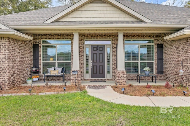 property entrance featuring a lawn, brick siding, a porch, and roof with shingles