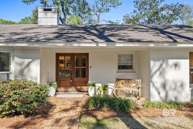 view of exterior entry featuring covered porch, a shingled roof, french doors, and brick siding