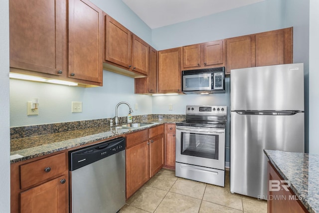 kitchen featuring a sink, dark stone countertops, stainless steel appliances, brown cabinetry, and light tile patterned floors