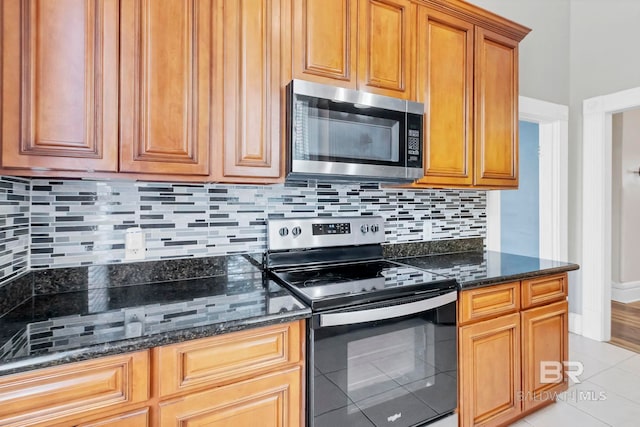 kitchen with appliances with stainless steel finishes, brown cabinetry, dark stone counters, light tile patterned floors, and tasteful backsplash