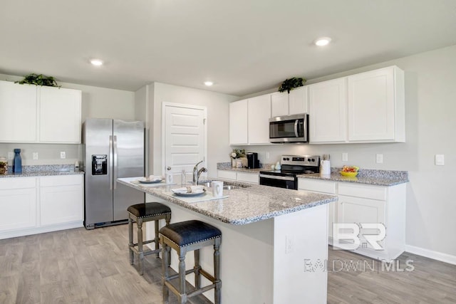 kitchen with an island with sink, light stone counters, appliances with stainless steel finishes, light wood-type flooring, and white cabinets