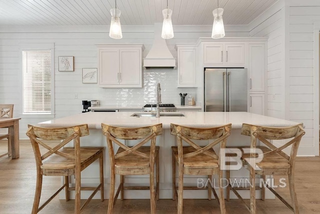 kitchen featuring custom exhaust hood, a kitchen island with sink, high quality fridge, and light countertops