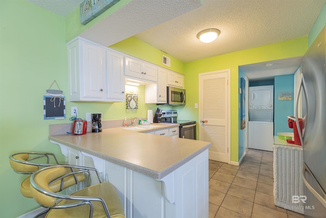 kitchen featuring white cabinets, kitchen peninsula, appliances with stainless steel finishes, stacked washer / dryer, and a textured ceiling