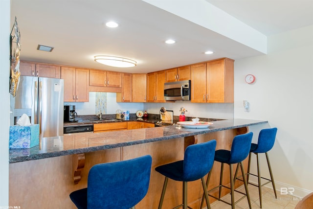 kitchen with dark stone countertops, light tile patterned floors, stainless steel appliances, sink, and a breakfast bar area