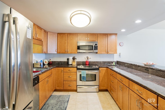 kitchen with dark stone counters, brown cabinets, appliances with stainless steel finishes, light tile patterned flooring, and a sink