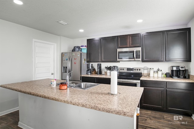 kitchen with a textured ceiling, a center island with sink, dark wood-type flooring, stainless steel appliances, and sink