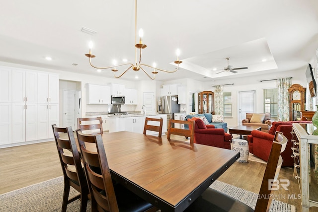 dining space with light wood-type flooring, ceiling fan with notable chandelier, a tray ceiling, and sink