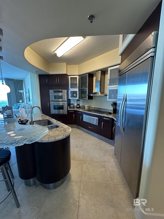 kitchen featuring sink, a breakfast bar area, light stone counters, hanging light fixtures, and wall chimney range hood