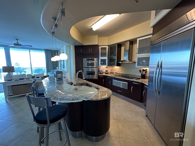 kitchen with pendant lighting, wall chimney range hood, sink, appliances with stainless steel finishes, and an island with sink