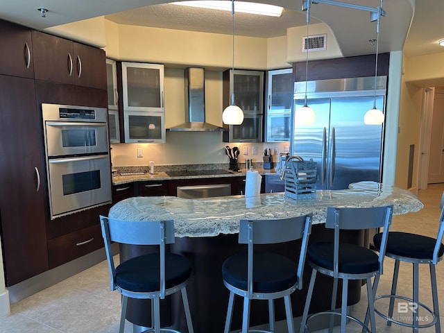 kitchen with dark brown cabinetry, decorative light fixtures, an island with sink, stainless steel appliances, and wall chimney range hood