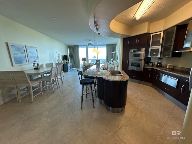 kitchen featuring double oven, sink, hanging light fixtures, light stone counters, and a center island with sink