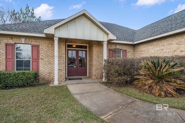 view of exterior entry featuring a shingled roof, a lawn, french doors, board and batten siding, and brick siding