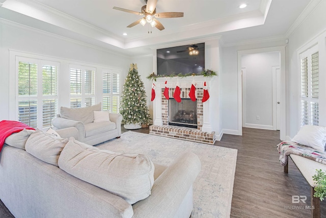 living room featuring a brick fireplace, dark hardwood / wood-style floors, ceiling fan, ornamental molding, and a tray ceiling