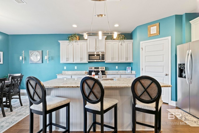 kitchen with appliances with stainless steel finishes, white cabinetry, a kitchen island with sink, light stone countertops, and decorative light fixtures