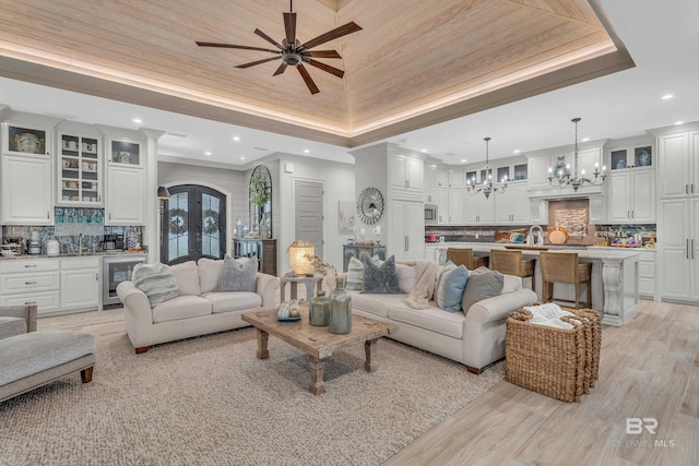 living room featuring indoor wet bar, light hardwood / wood-style floors, a raised ceiling, ceiling fan with notable chandelier, and beverage cooler