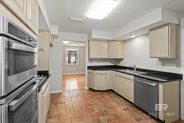 kitchen with a sink, brick floor, cream cabinets, stainless steel appliances, and baseboards