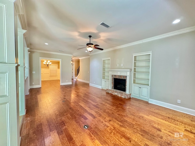 unfurnished living room with wood-type flooring, ornamental molding, a brick fireplace, built in features, and ceiling fan with notable chandelier