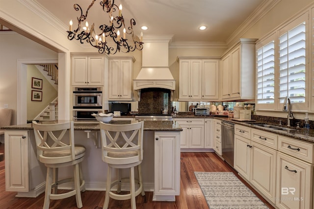 kitchen featuring appliances with stainless steel finishes, premium range hood, sink, dark stone countertops, and hanging light fixtures