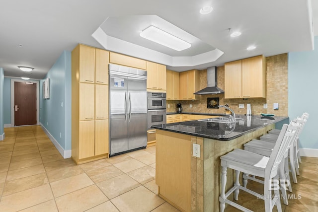 kitchen featuring stainless steel appliances, kitchen peninsula, tasteful backsplash, wall chimney exhaust hood, and dark stone countertops