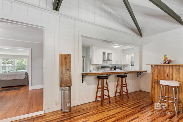 kitchen with kitchen peninsula, vaulted ceiling, wall chimney exhaust hood, a kitchen bar, and white cabinetry