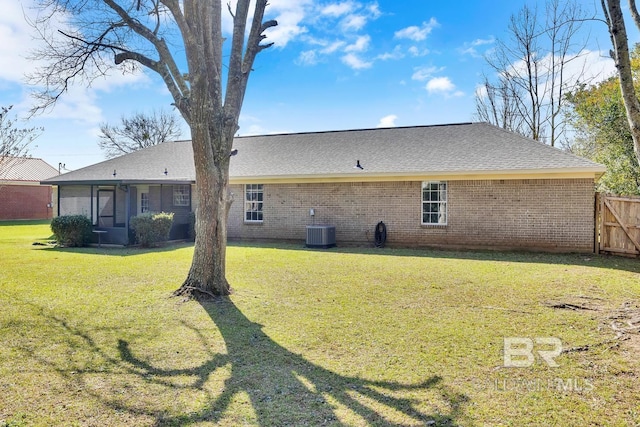 rear view of property with brick siding, fence, roof with shingles, central AC unit, and a yard