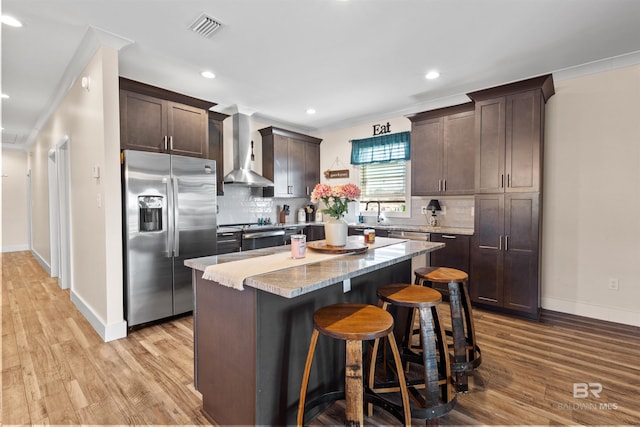 kitchen featuring visible vents, wall chimney exhaust hood, a kitchen island, appliances with stainless steel finishes, and dark brown cabinets