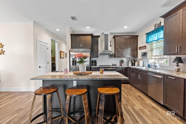 kitchen featuring dark brown cabinetry, wall chimney exhaust hood, appliances with stainless steel finishes, a center island, and a sink