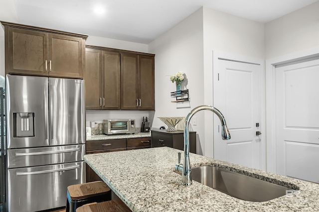 kitchen featuring a sink, stainless steel refrigerator with ice dispenser, dark brown cabinets, and light stone countertops