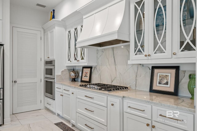 kitchen with appliances with stainless steel finishes, tasteful backsplash, light stone counters, custom exhaust hood, and white cabinetry