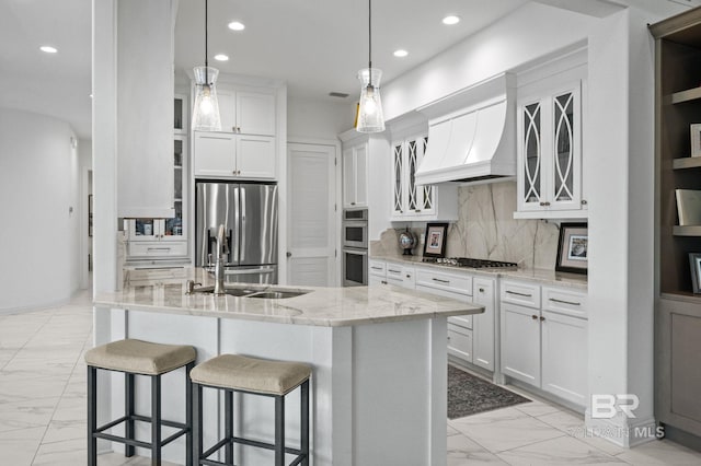 kitchen featuring appliances with stainless steel finishes, custom range hood, a center island with sink, decorative light fixtures, and white cabinets