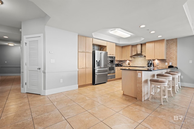 kitchen featuring a kitchen breakfast bar, wall chimney range hood, light brown cabinetry, kitchen peninsula, and stainless steel appliances