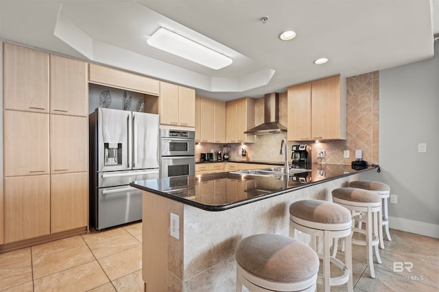 kitchen featuring light brown cabinets, dark stone counters, wall chimney exhaust hood, kitchen peninsula, and stainless steel appliances